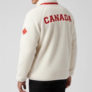 May include: Cream-colored fleece jacket with a red "CANADA" graphic on the back and a red maple leaf emblem. The jacket has a red trim around the collar and a quarter-zip closure. The model is wearing black pants.