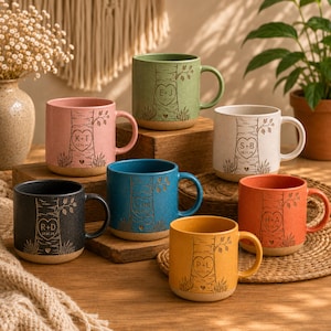Op de afbeelding: Een verzameling keramische mokken in verschillende kleuren, waaronder roze, groen, blauw, oranje, geel, zwart en wit. Elke mok heeft een boomontwerp met initialen en data gegraveerd. De mokken hebben een rustieke, handgemaakte uitstraling.