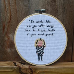 May include: A cross-stitch embroidery in a round wooden hoop. The text reads "Be careful John, lest you suffer vertigo from the dizzying heights of your moral ground." A cartoon figure is stitched below the text.