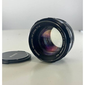 May include: A black Konica Hexanon AR 57mm F1.4 camera lens with a lens cap. The lens is made in Japan and has a serial number. The lens is on a white surface.