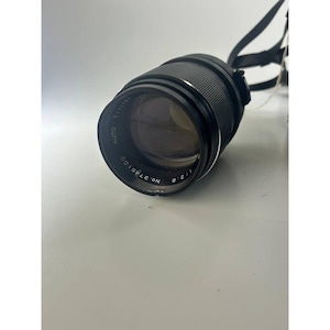 May include: A black camera lens with the text "AUTO VIVITAR" and "NO. 3785100" printed on the lens. The lens has a black strap attached. The lens is lying on a white surface.