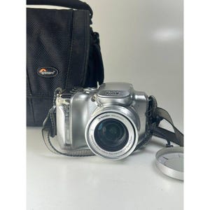 以下が含まれることがあります： 黒いレンズと黒いカメラバッグが付いた、シルバーのKodakデジタルカメラ。カメラにはストラップとレンズキャップが付いています。カメラバッグには「lowepro」の文字が入っています。