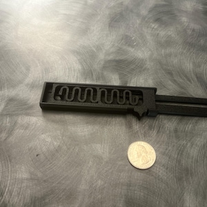 May include: A black, 3D-printed key with a unique internal design, next to a U.S. quarter for size comparison. The key has a rectangular head with a wavy pattern and a long, flat handle. The background is a brushed metal surface.
