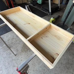 May include: A rectangular, unfinished wooden box with a raised edge, supported by two metal sawhorses. The box is divided into two sections by a central divider. The wood grain is visible, and the box appears to be newly constructed.