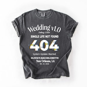Peut inclure: T-shirt gris foncé avec texte blanc : "Wedding v1.0 Debug Crew SINGLE LIFE NOT FOUND 404 System Update: Married OLIVIA'S BACHELORETTE New Orleans, LA 08.15.2026". Les manches sont retroussées.