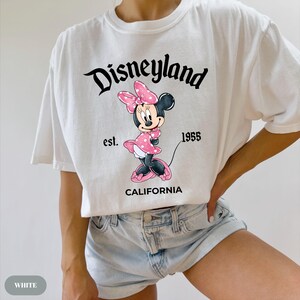 May include: White t-shirt featuring a graphic of Minnie Mouse in a pink polka-dot dress and bow. The shirt has the text "Disneyland" above the character, with "est. 1955" and "CALIFORNIA" below.