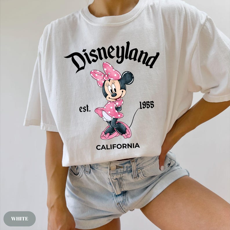 May include: White t-shirt featuring a graphic of Minnie Mouse in a pink polka-dot dress and bow. The shirt has the text "Disneyland" above the character, with "est. 1955" and "CALIFORNIA" below.