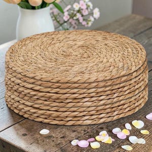 May include: A stack of woven, circular placemats made from natural fibres. The placemats are a light tan colour and have a spiral design in the centre. The stack is on a wooden table with flower petals scattered around.