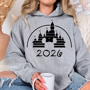 Puede incluir: Sudadera con capucha gris jaspeado con un dise&ntilde;o gr&aacute;fico negro de un castillo que incorpora una silueta de Mickey Mouse y el a&ntilde;o "2026". La sudadera tiene un bolsillo delantero y una capucha con cord&oacute;n.