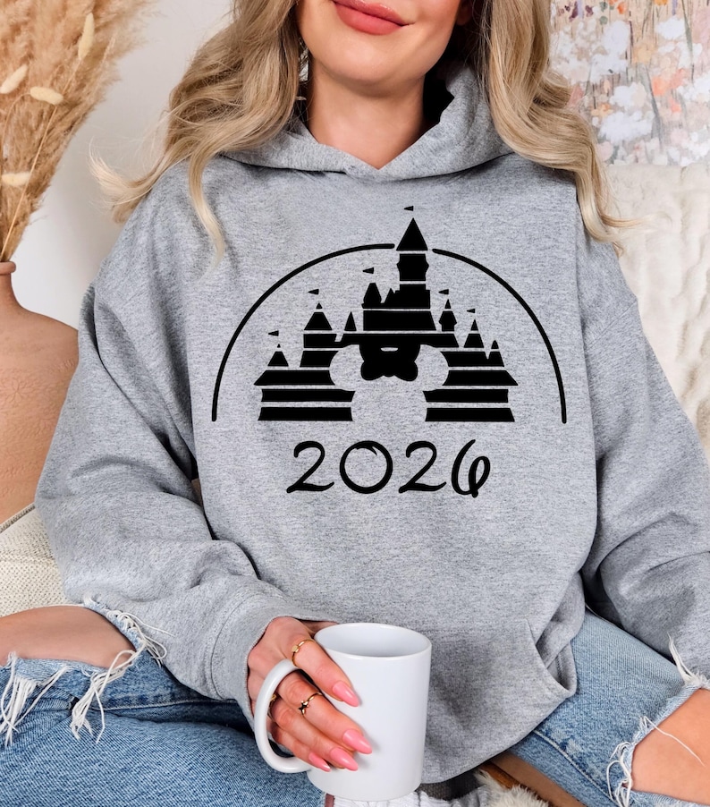 Puede incluir: Sudadera con capucha gris jaspeado con un dise&ntilde;o gr&aacute;fico negro de un castillo que incorpora una silueta de Mickey Mouse y el a&ntilde;o "2026". La sudadera tiene un bolsillo delantero y una capucha con cord&oacute;n.