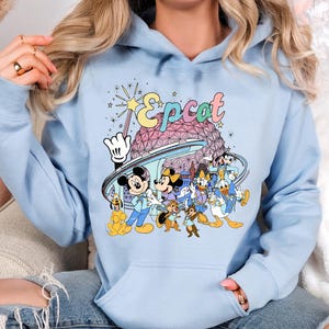 May include: Light blue hoodie featuring a colorful graphic of Disney characters, including Mickey and Minnie Mouse, Donald Duck, and Goofy, with the word "Epcot" above them. The design includes the Epcot ball and other park elements.
