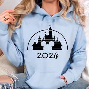 Puede incluir: Sudadera con capucha azul claro con un dise&ntilde;o negro de un castillo con orejas de Mickey Mouse y el a&ntilde;o 2026. La sudadera tiene un bolsillo delantero y una capucha con cord&oacute;n. Las mangas son largas.