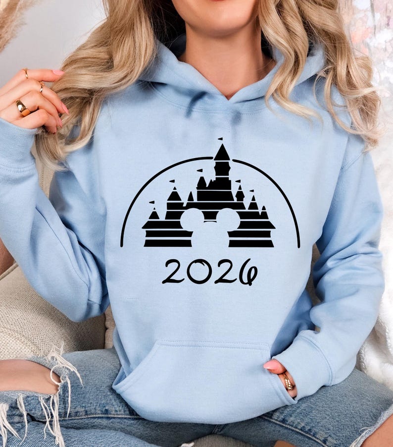 Puede incluir: Sudadera con capucha azul claro con un dise&ntilde;o negro de un castillo con orejas de Mickey Mouse y el a&ntilde;o 2026. La sudadera tiene un bolsillo delantero y una capucha con cord&oacute;n. Las mangas son largas.