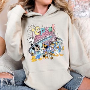 May include: A beige hoodie featuring a colorful graphic of the Epcot logo and various Disney characters. The design includes Mickey Mouse, Minnie Mouse, Donald Duck, and others, with a stylized Spaceship Earth in the background.