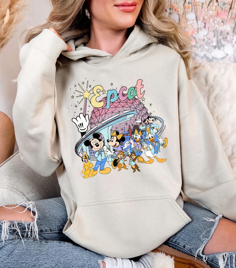 May include: A beige hoodie featuring a colorful graphic of the Epcot logo and various Disney characters. The design includes Mickey Mouse, Minnie Mouse, Donald Duck, and others, with a stylized Spaceship Earth in the background.