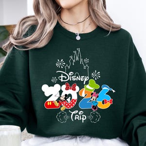 Pu&ograve; includere: Felpa verde scuro con la scritta "Disney 2023 Trip" e illustrazioni di Topolino e Minnie. Il design include una silhouette di castello e numeri colorati. Un top comodo e casual.