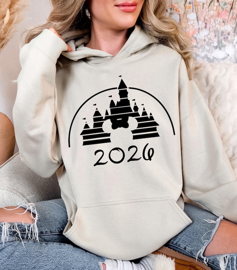 Puede incluir: Sudadera con capucha beige con un dise&ntilde;o gr&aacute;fico negro de un castillo y el a&ntilde;o 2026 impreso debajo. La sudadera tiene un bolsillo delantero y una capucha con cord&oacute;n. El dise&ntilde;o incorpora una cabeza de Mickey Mouse.