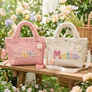 Puede incluir: Dos bolsos tote de pana con asas y correas para el hombro. Uno es rosa con el nombre "Luna" en letras pastel, el otro es blanco roto con "Mattia". Ambos tienen adornos de flores. Están sobre un banco de madera.