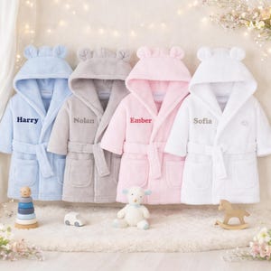 May include: Four plush baby bathrobes in pastel colors: light blue, gray, pink, and white. Each robe has a hood with bear ears and a matching belt. Names are embroidered on the front: Harry, Nolan, Ember, and Sofia. A wooden toy stacker, car, lamb, and rocking horse are in front.