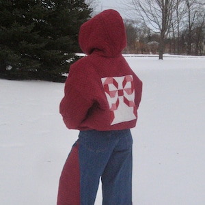 May include: A person wearing a red hooded jacket with a white and red quilt block design on the back, paired with blue jeans featuring red side panels. The person is standing in the snow.