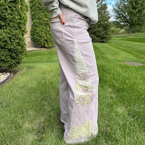 May include: Light purple denim pants featuring a vertical side panel of quilted fabric. The quilted section displays a patchwork design with floral patterns in shades of green, white, and purple. The pants have a standard five-pocket construction.