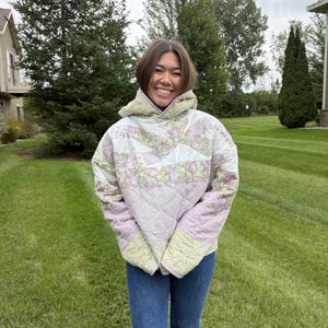 Puede incluir: Una chaqueta acolchada con capucha que presenta un diseño de patchwork con patrones florales y botánicos en tonos verde salvia, lavanda y blanco. La prenda tiene mangas largas, una capucha ancha y una silueta cuadrada con costuras en rombos.
