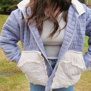 May include: A light blue quilted jacket with white accents on the collar and pockets. The jacket has a diamond pattern and is worn open, revealing a gray shirt and blue jeans. The pockets have a ruffled edge.