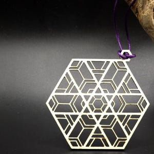 Puede incluir: Colgante geométrico de madera clara cortada con láser, con un diseño hexagonal complejo. Cuelga de un cordón morado. El fondo es negro con un trozo de madera flotante en la esquina superior derecha.
