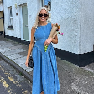 Può includere: Abito senza maniche in denim blu chiaro con gonna svasata. L'abito presenta un corpetto aderente e uno scollo tondo. La persona indossa occhiali da sole neri e sandali neri con fibbie dorate. Tiene in mano un mazzo di fiori.