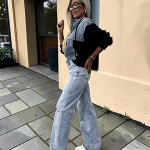 May include: A woman wearing a denim and black color block jacket, light wash jeans, white sneakers, and glasses. The jacket has a hood and the jeans are wide-legged. The background is a building with a beige exterior and a paved walkway.