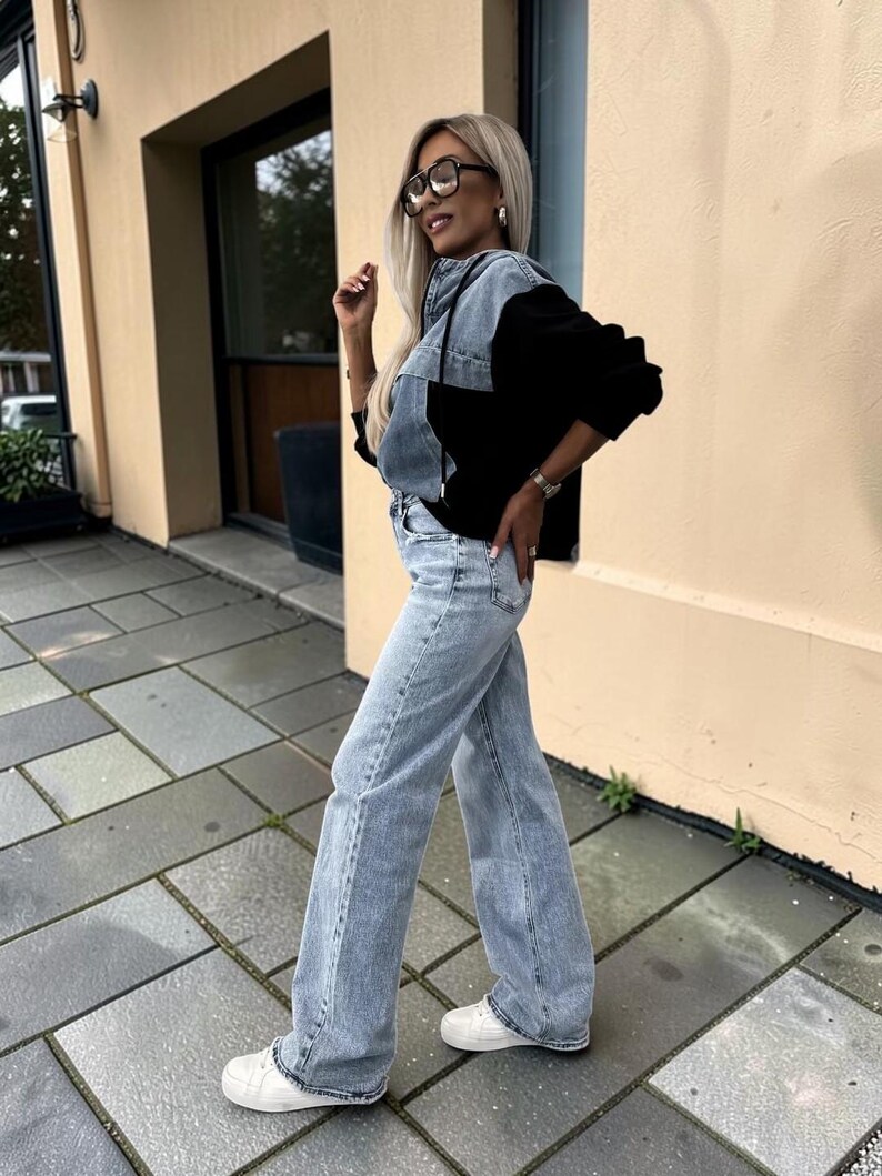 May include: A woman wearing a denim and black color block jacket, light wash jeans, white sneakers, and glasses. The jacket has a hood and the jeans are wide-legged. The background is a building with a beige exterior and a paved walkway.