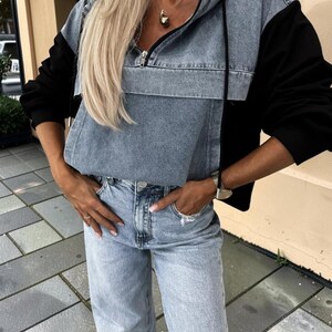 May include: A denim and black hooded sweatshirt with a front zipper and a front pocket. The model is wearing light blue denim jeans. The outfit is casual and stylish.