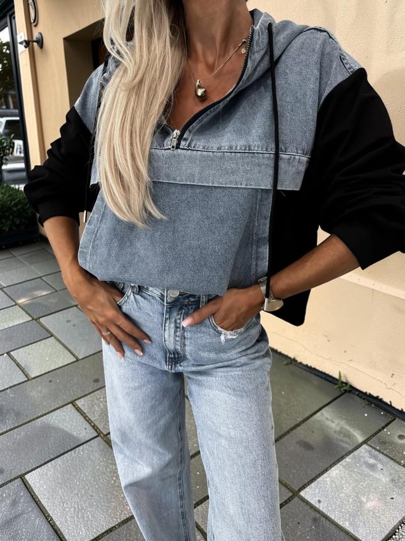 May include: A denim and black hooded sweatshirt with a front zipper and a front pocket. The model is wearing light blue denim jeans. The outfit is casual and stylish.