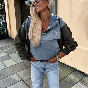 May include: A denim and olive green hooded jacket with a front zipper, paired with light blue jeans. The jacket has a color-block design, with denim on the upper body and olive green sleeves. The jeans are a light wash.
