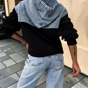 May include: Black hooded jacket with a light blue denim hood and shoulders. The jacket has long black sleeves and is paired with light blue denim jeans. The model is wearing black sunglasses.