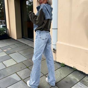 May include: A light-wash denim jacket with a dark olive green panel is paired with light-wash denim jeans and white sneakers. The jacket has a hood and the jeans have a wide leg. The outfit is worn outdoors.