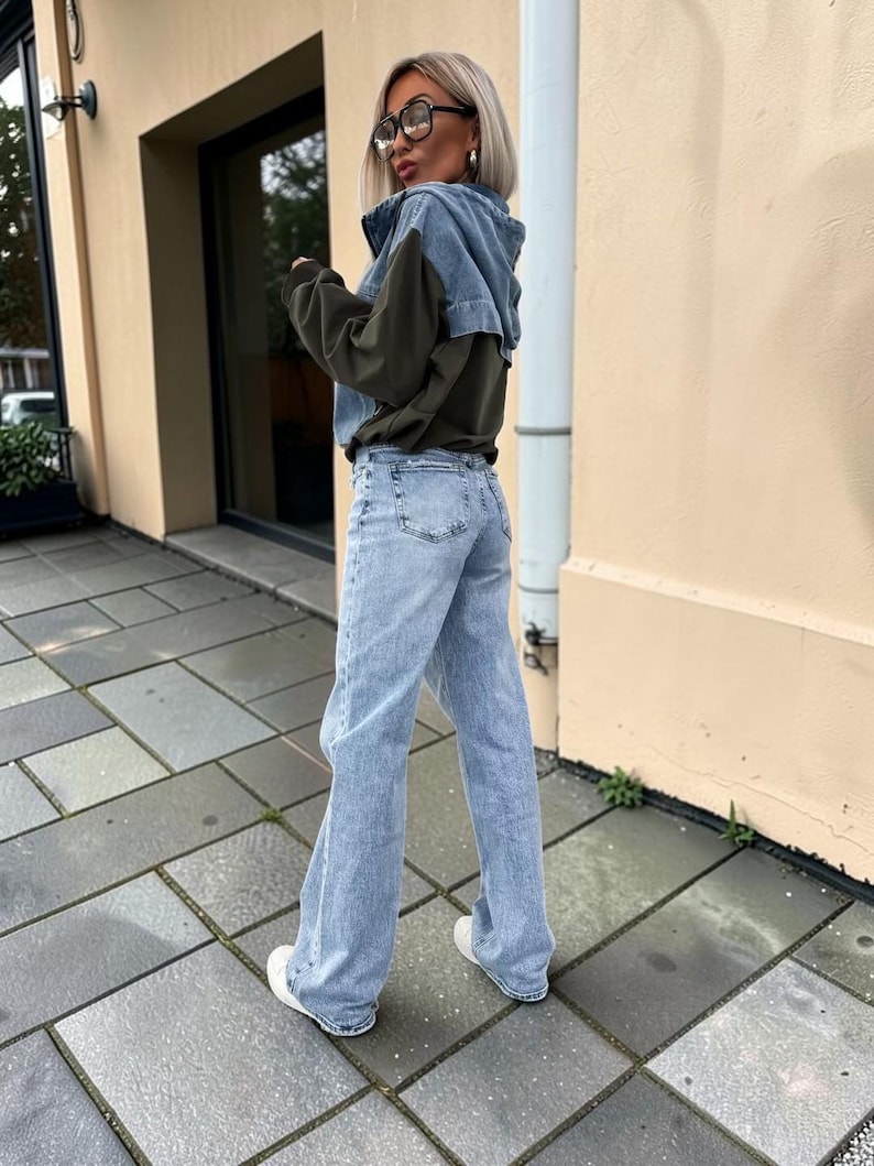 May include: A light-wash denim jacket with a dark olive green panel is paired with light-wash denim jeans and white sneakers. The jacket has a hood and the jeans have a wide leg. The outfit is worn outdoors.