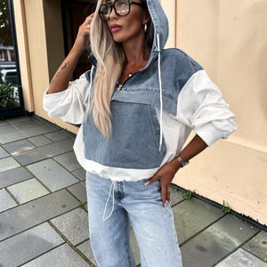 May include: A hooded denim and white jacket with a front pocket and drawstring hem. The jacket is paired with light wash denim jeans. The model is wearing large black-framed glasses.