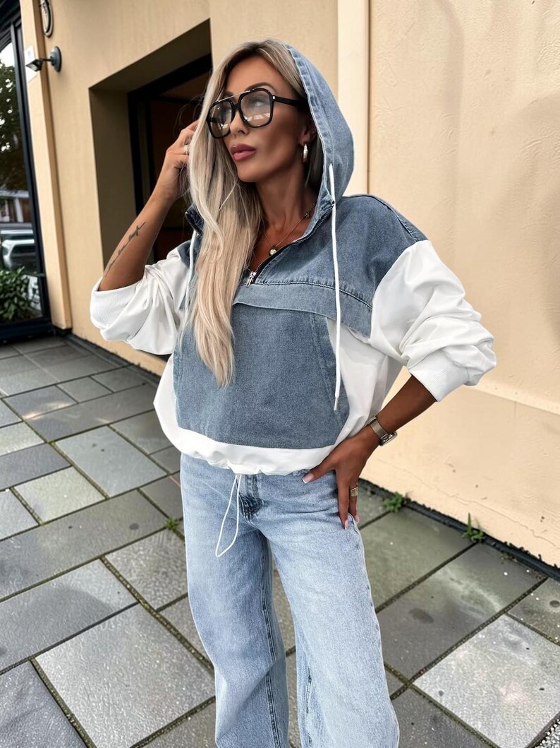May include: A hooded denim and white jacket with a front pocket and drawstring hem. The jacket is paired with light wash denim jeans. The model is wearing large black-framed glasses.