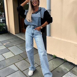 May include: A light blue denim hoodie with black sleeves and light blue jeans. The outfit is paired with white sneakers. The model is wearing black-framed glasses and a watch.
