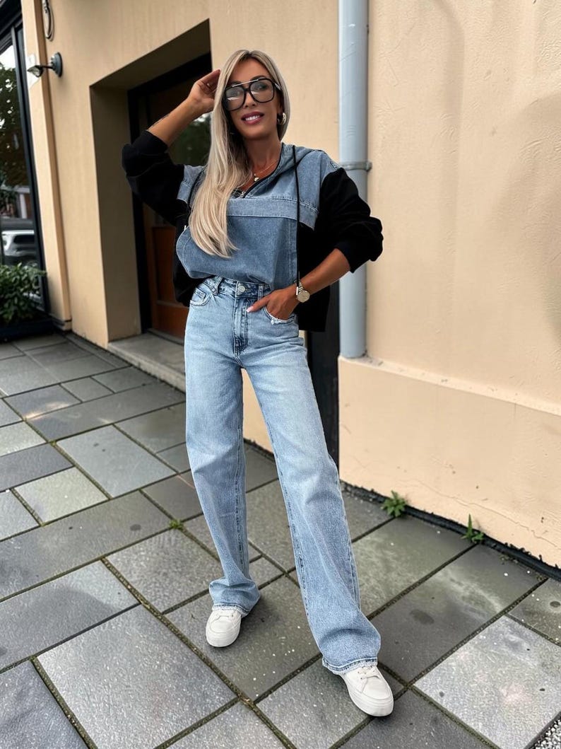 May include: A light blue denim hoodie with black sleeves and light blue jeans. The outfit is paired with white sneakers. The model is wearing black-framed glasses and a watch.