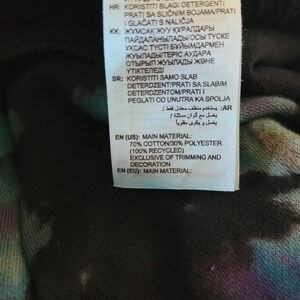 May include: A close-up of a clothing tag with text in multiple languages. The tag lists the main material as 70% cotton and 30% polyester, with 100% recycled content. The tag also includes washing instructions.