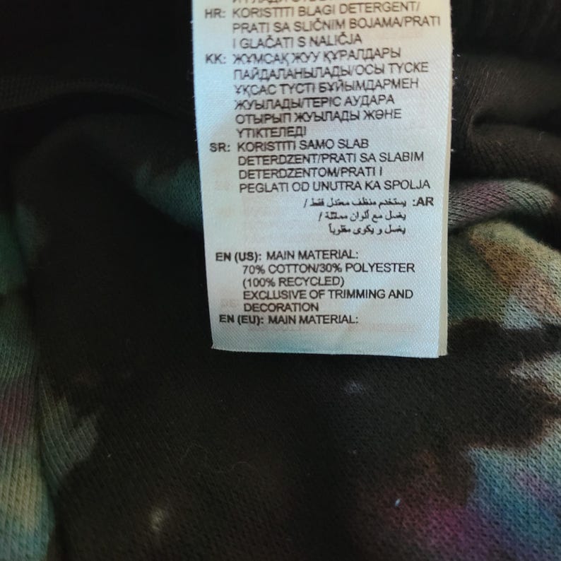 May include: A close-up of a clothing tag with text in multiple languages. The tag lists the main material as 70% cotton and 30% polyester, with 100% recycled content. The tag also includes washing instructions.