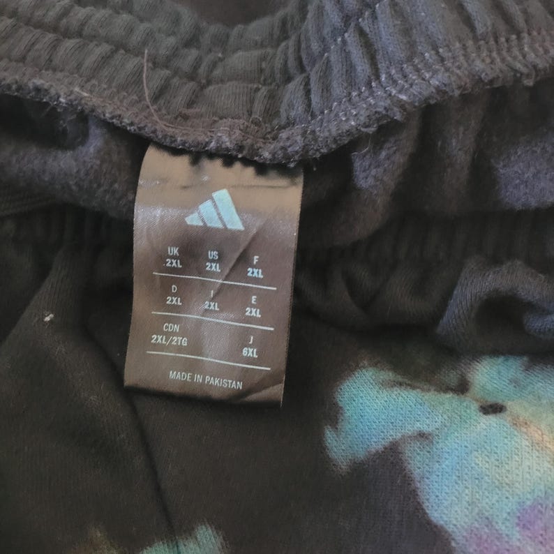 May include: Close-up of a black garment tag with the Adidas logo and size information. The tag indicates sizes for various regions, including US 2XL. The tag is labeled "MADE IN PAKISTAN".