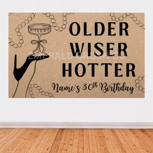 May include: A brown sign with the words "OLDER WISER HOTTER" in black text, with a champagne glass illustration. The sign also includes the text "Name's 30th Birthday". Two black balloons are on either side of the sign.