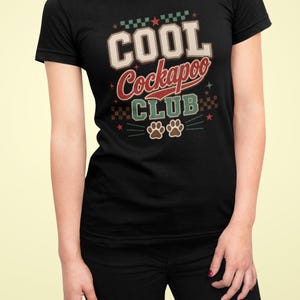 Könnte beinhalten: Ein schwarzes Kurzarm-T-Shirt mit einem Grafikdesign, das den Text "COOL Cockapoo CLUB" in einer College-Schriftart zeigt. Das Design enthält rote, weiße und grüne Akzente, ein Karomuster sowie zwei braune Pfotenabdruck-Symbole.