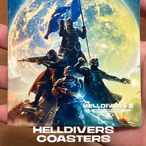 May include: Square coaster featuring a sci-fi illustration of soldiers with weapons, a flag, and a planet in the background. The coaster has the text "HELLDIVERS COASTERS" in white letters.