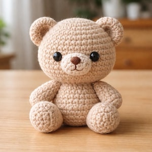 Könnte beinhalten: Ein handgefertigter, hellbrauner Häkelteddybär mit schwarzen Sicherheitsaugen und einer braunen Nase. Der Bär hat einen lächelnden Ausdruck und sitzt auf einer Holzoberfläche. Die Arme und Beine des Bären sind ebenfalls gehäkelt.