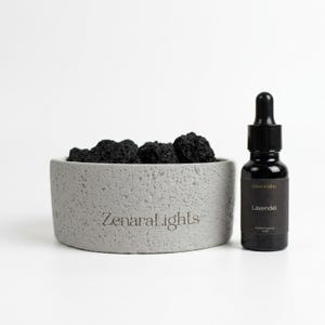 Könnte beinhalten: Eine hellgraue, texturierte Betonschale gefüllt mit dunklen, porösen Lavasteinen, mit dem Schriftzug ZenaraLights auf der Vorderseite. Daneben steht eine kleine, dunkelbraune Glastropfflasche mit dem Etikett ZenaraLights Lavendel Aroma Essence.