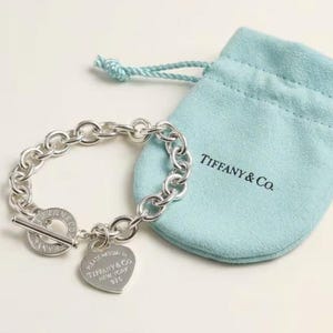 May include: A silver chain bracelet with a heart-shaped charm and a toggle clasp. The charm is engraved with text. A light blue drawstring pouch with the text "TIFFANY & Co." is next to the bracelet.
